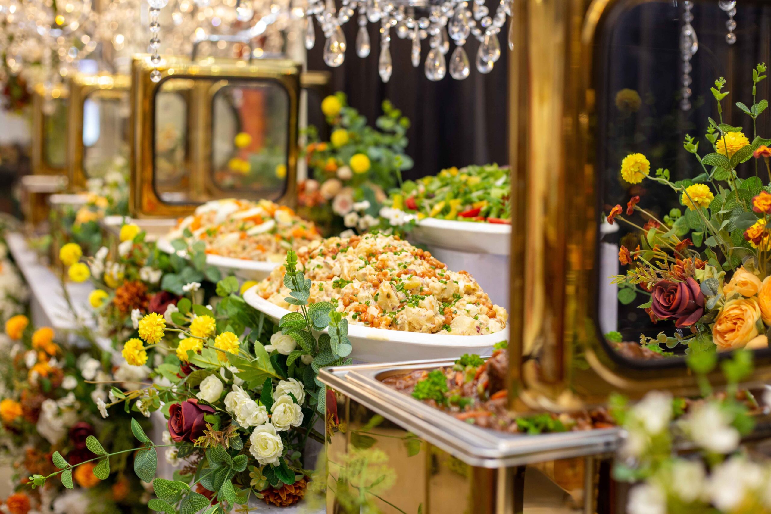 Buffet table decorated with flowers and serving dishes filled with colorful food at an event.